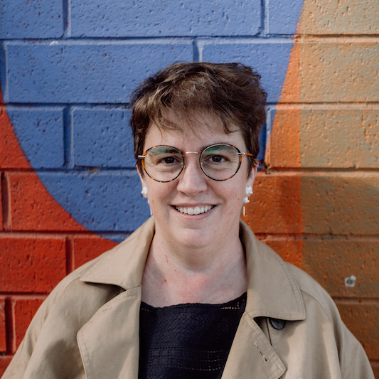 Image of Rachel Amies wearing glasses, white cat earrings, a dark blue top and a light jacket. She's leaning against a brick wall that's been painted colourfully in blue, red, orange and yellow.