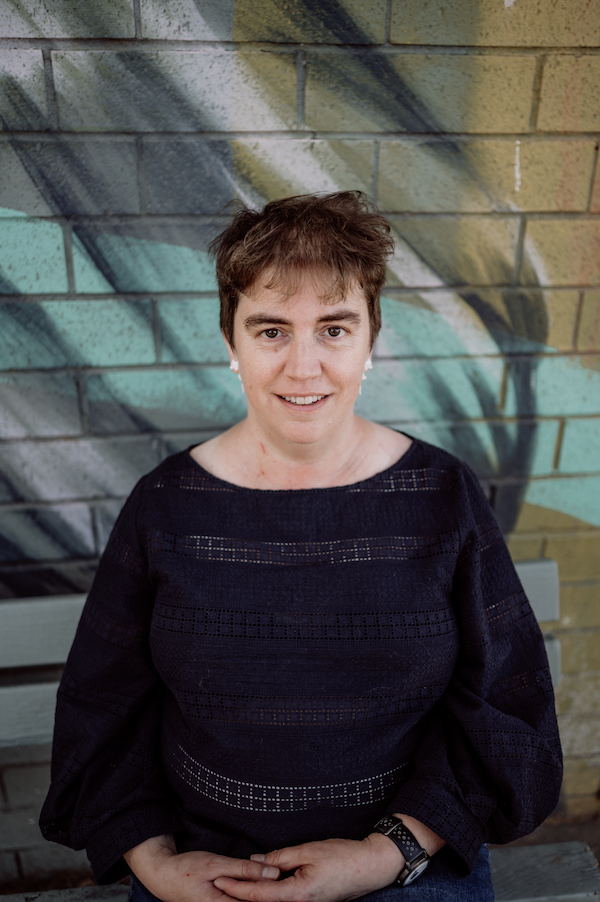 Image of Rachel Amies who's wearing a dark blue top, white cat earrings and sitting down at a bench. The bench backs onto a brick wall that is painted colourfully.