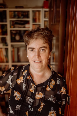 Rachel Amies wearing a black shirt with white cats and ginger cats on it. She's wearing cat earrings and is standing in the doorway with bookshelves in the background.